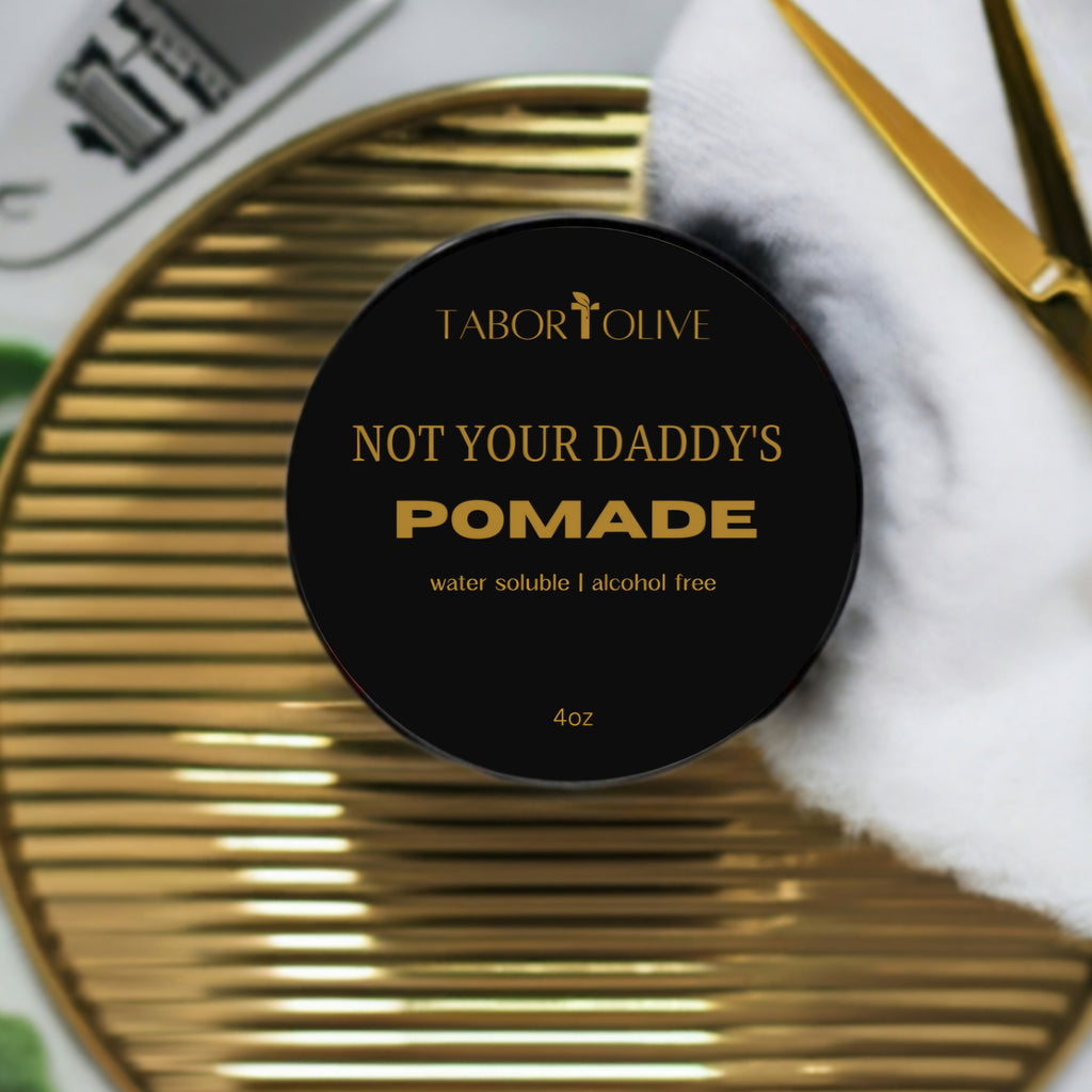 Black jar flat lay on gold surface next to white towel and gold scissors. The blackjar has gold text that says, "not your Daddys pomade. Water Soluble. Alcohol free."