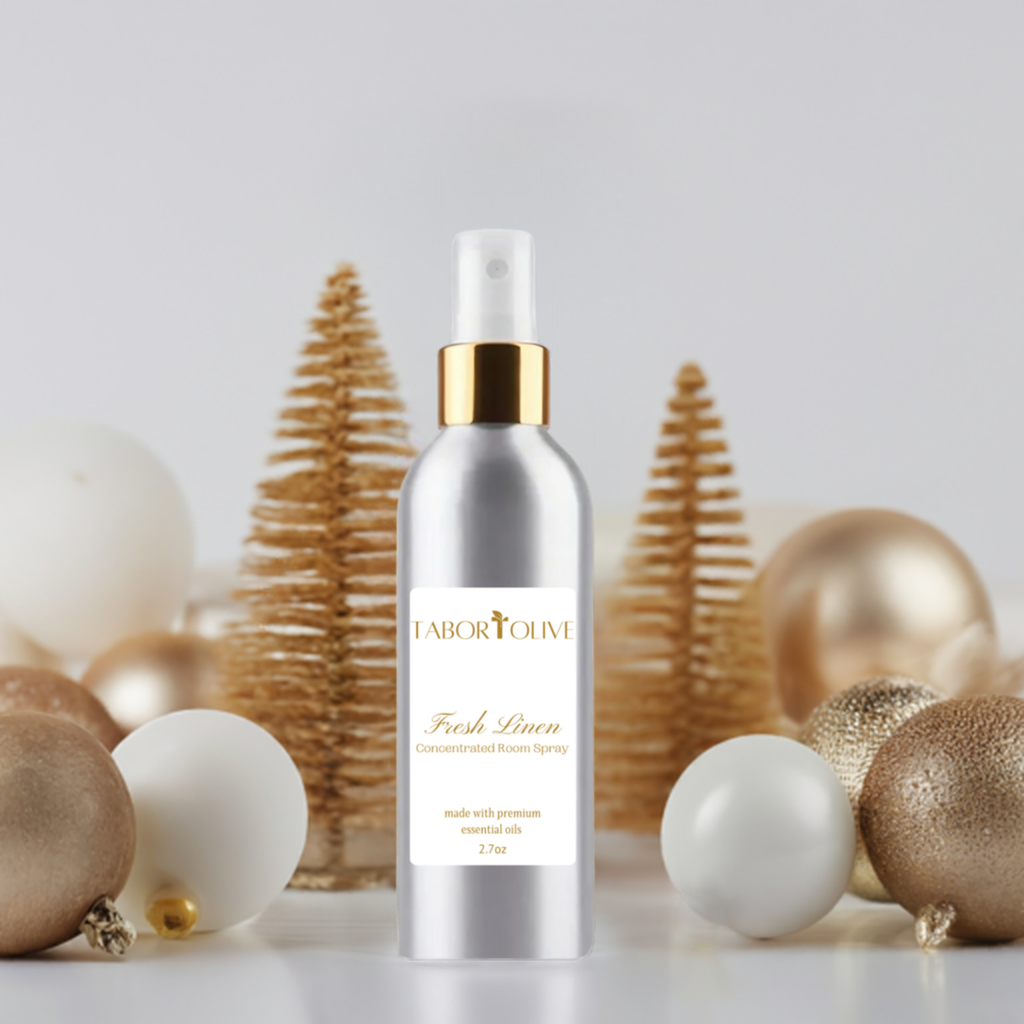 Aluminum spray bottle with gold cap and white label with gold text that says, "fresh linen concentrated room sprays. Made with premium essential oils." Sitting on marble platform next to gold Christmas decorations.