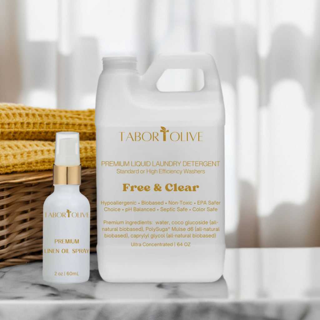 White detergent bottle with gold text that says, "free and clear" next to white linen oil bottle on a marble platform next to rust orange colored towels in woven basket.