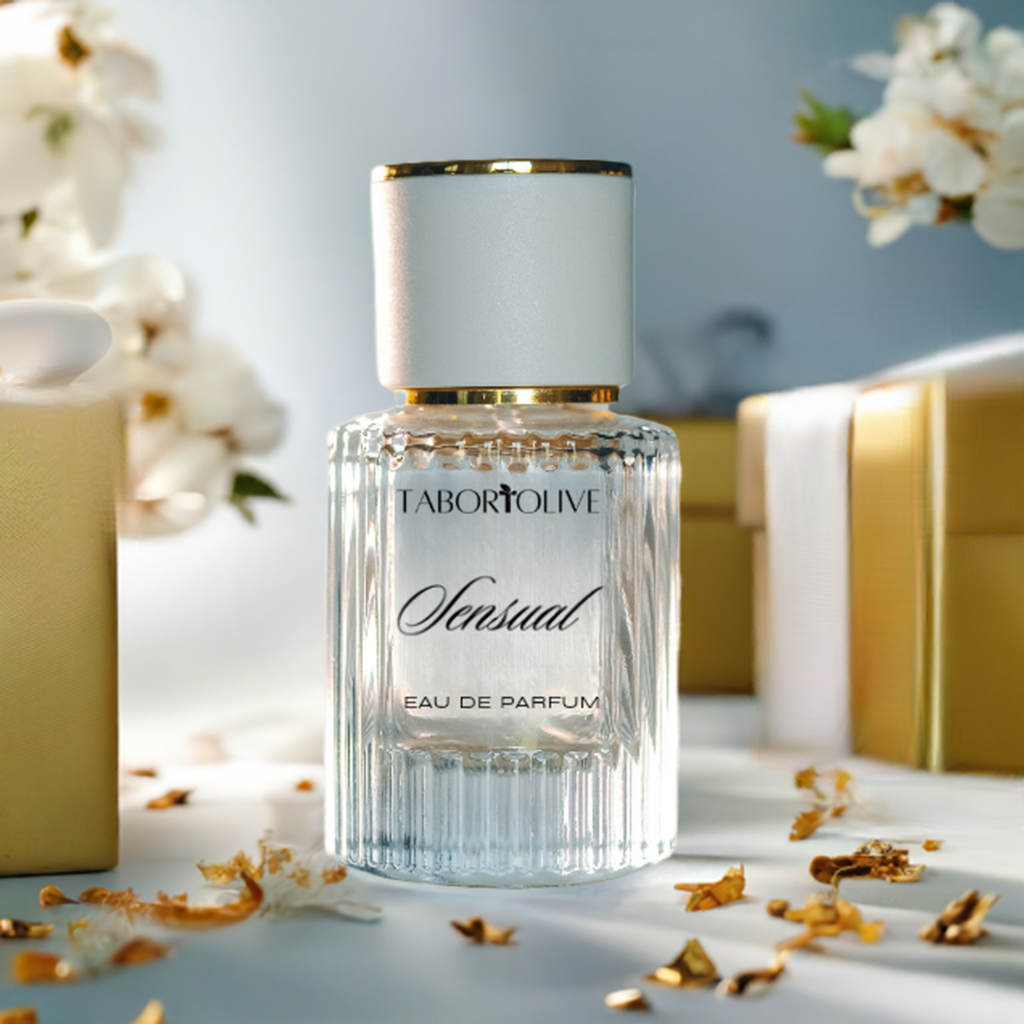 Clear textured glass perfume bottle next to gold gift boxes and white flowering branches with text on the bottle that says, "sensual eau de parfum"